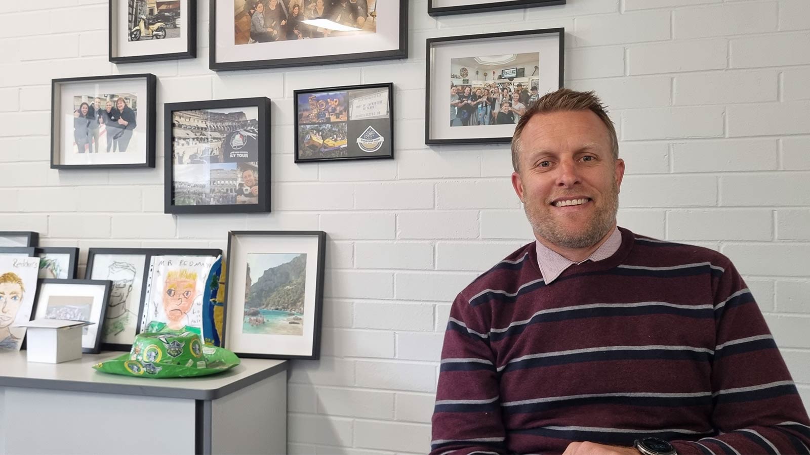 Nathan Redmayne has dark blond hair and a light bear. He is sitting behind his desk in front of a white brick wall with framed pictures on it.