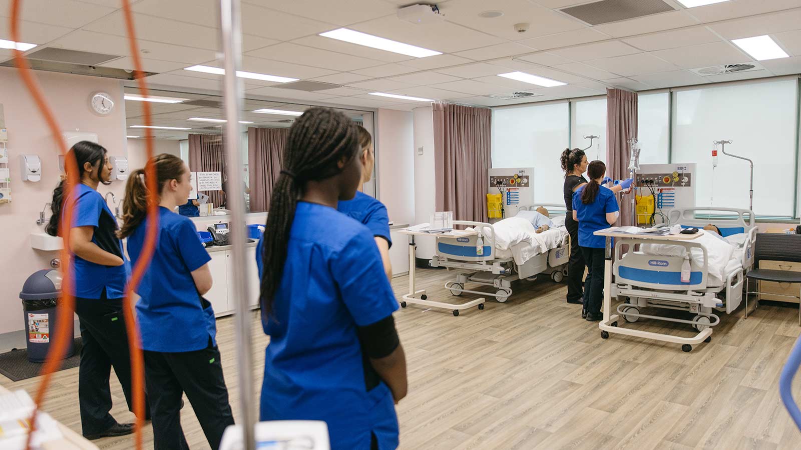 A group of nursing students are in a simulation lab at UOW watching a demonstration on a dummy patient. They are wearing dark blue UOW scrubs.