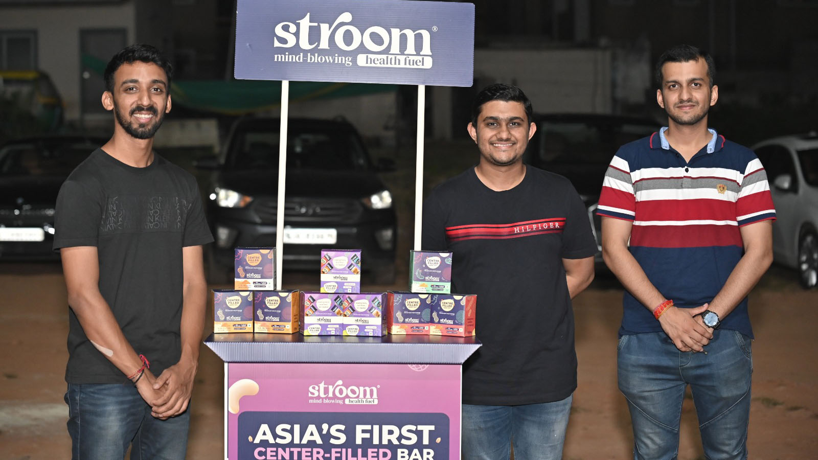 Three men are standing at a purple 'Stroom' booth, advertising energy bars