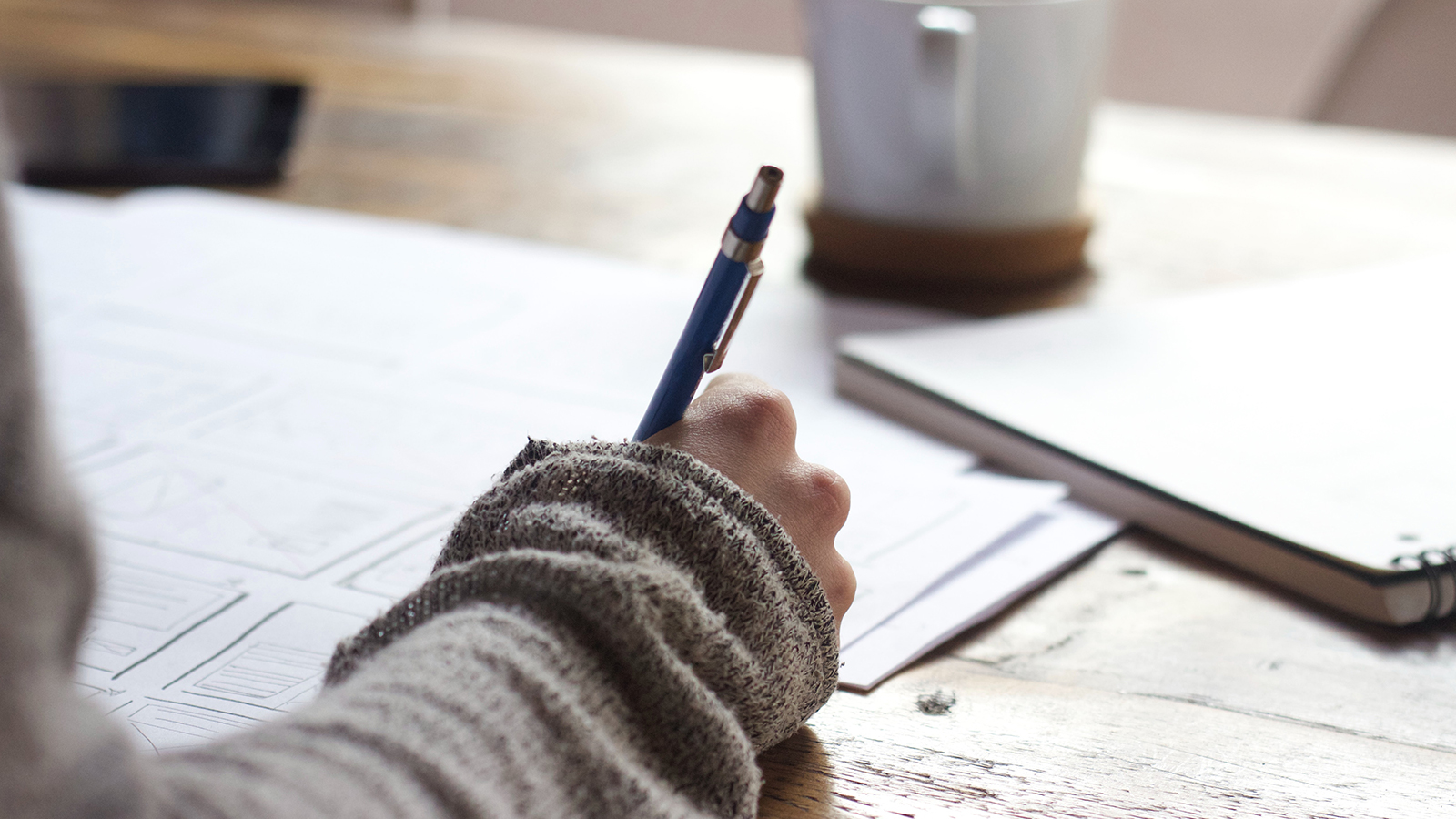 A hand holding a pen, writing in a notebook. They have a grey sleeve and a coffee cup on the table.