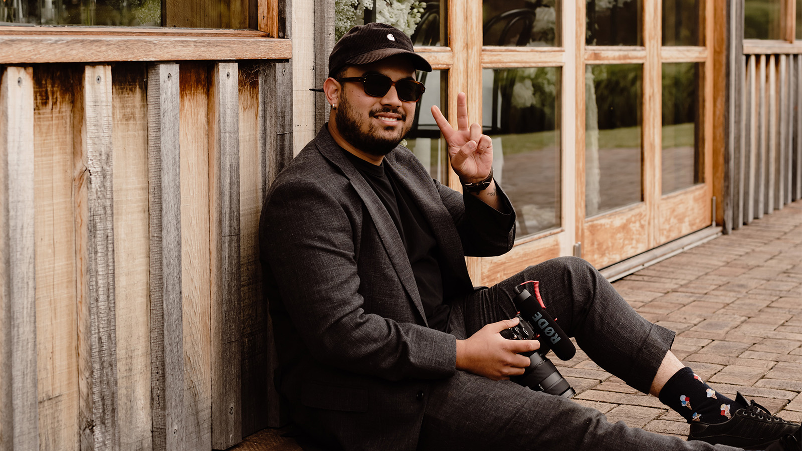 A man is sitting down against a rustic barn, holding a video camera and doing a peace sign with his hands, smiling