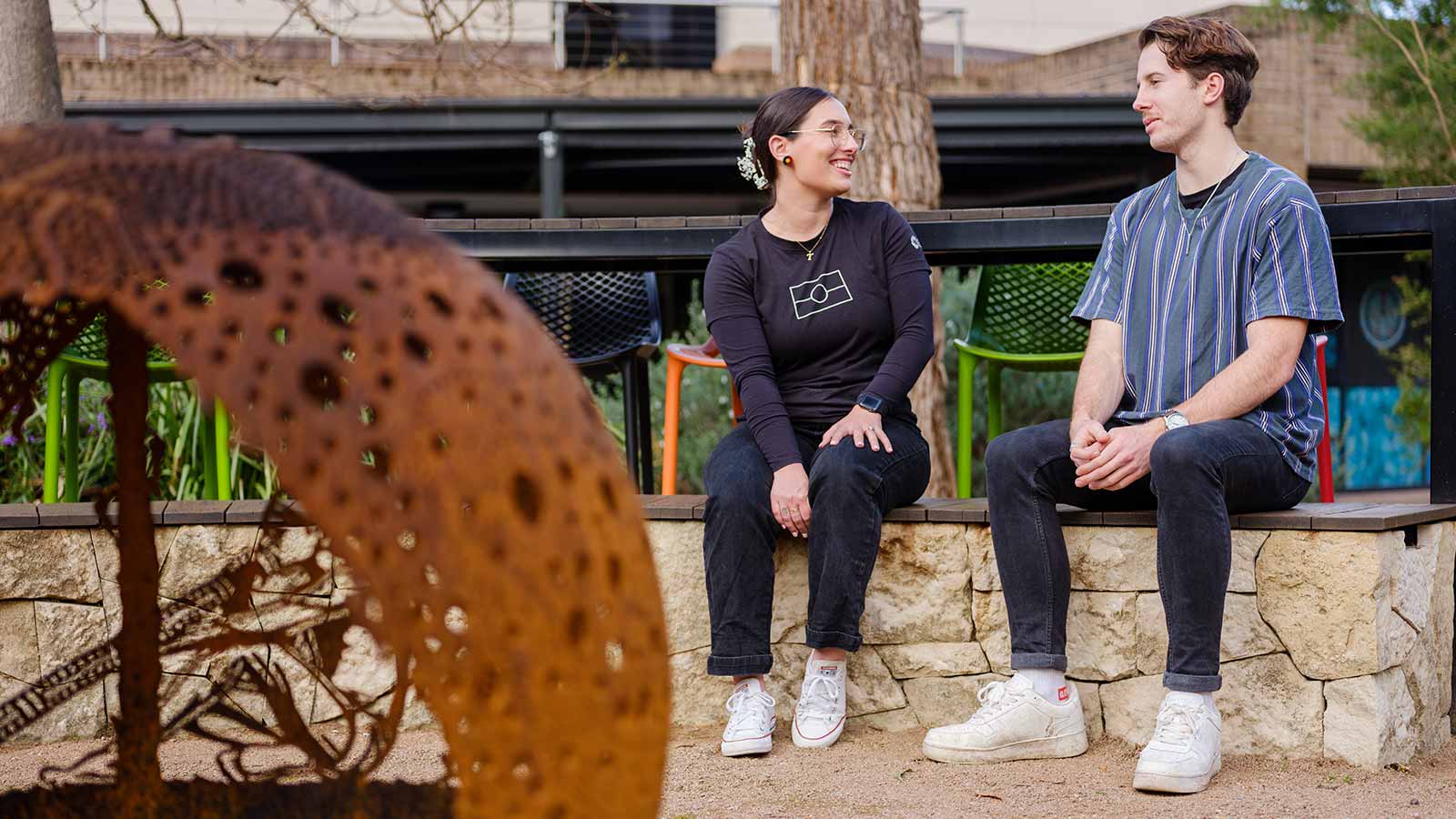 A woman and a man are sitting on a stone bench seat outside UOW Woolyungah Indigenous Centre. They are at behind a brown, spherical fire pit.