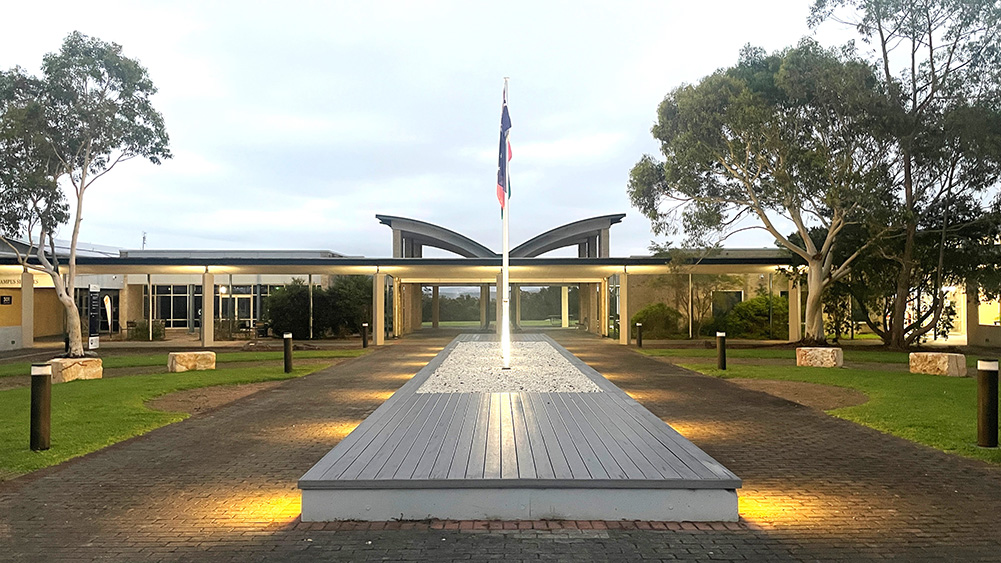 A serene courtyard featuring a central flagpole, illuminated pathway, greenery, and modern buildings under a cloudy sky.