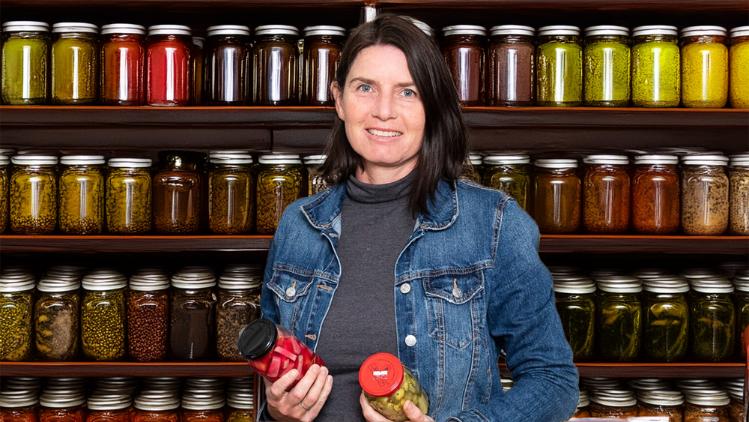 A lady is standing in front of shelves full of jars. She holds two jars in each hand.