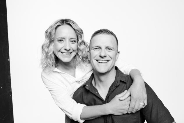 Black and white photo of a couple. The man appears to be sitting, with the fair haired woman standing behind him with her arms around his shoulders and her hands meeting in the middle.  Both are smiling at the camera.