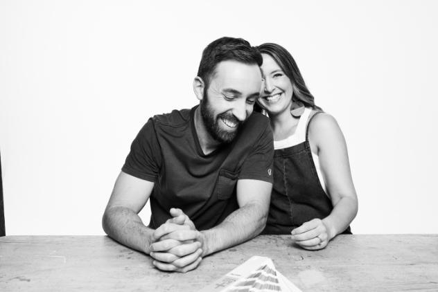 This is a black and white photo of a young couple sitting at a table with a colour chart in front of them. Matt has both hands resting on the table and is smiling looking down. Claire is slightly behind him and is smiling towards the camera.