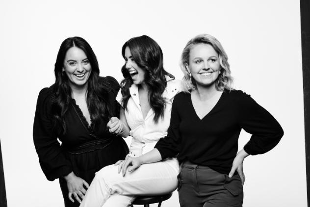 This photo is in black and white. Image of three young women, two standing and the woman in the middle is sitting on a stool. The woman on the left and the middle both have long dark hair and are laughing. The woman on the right has light hair and is smiling and laughing looking off to the side.