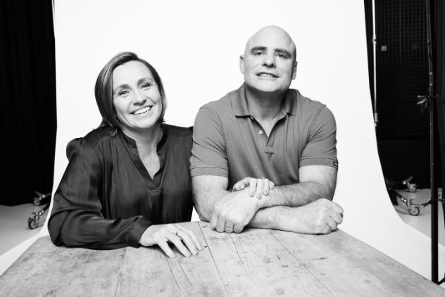 Black and white photo of a couple sitting together at a table. The woman has short hair and has a hand through the man's crossed arms. The man is bald and has his arms crossed.