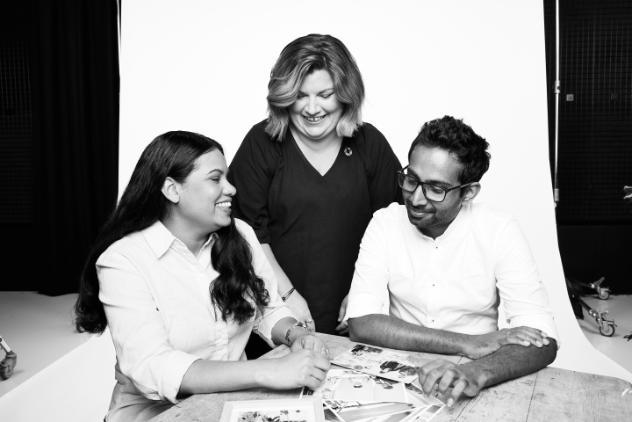 Black and white photo. Three people are together at a table with flyers in front of them. There is two females and one male. The first female on the left is sitting smiling at the man on the right. Sitting next to her is a man with glasses who is smiling at the flyers. Standing behind the middle of them is another woman who is also smiling and looking down at the camera.