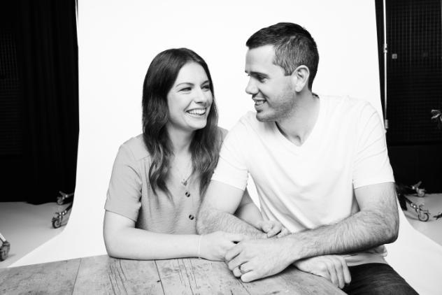 Black and white photo of a young couple sitting at a table. The pair are looking at each other smiling with their arms linked.