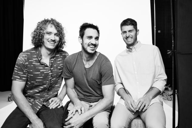 This is a black and white photo of three males sitting together. All of the men are laughing and looking at the camera except the middle man who is looking to the side.  The man on the right has shoulder length curly hair whilst the other men have short dark hair.