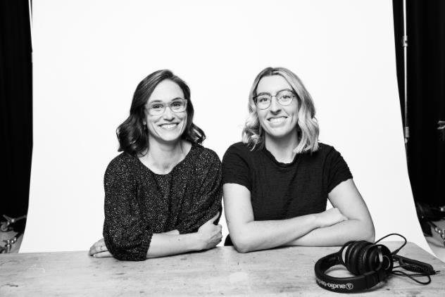 This is a black and white photo of two women sitting at a table. Both woman are smiling at the camera if their arms folded on the table. There is a pair of headphones on the bottom right hand corner of the table.