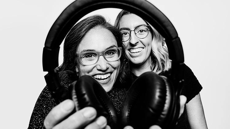 Lizzie Jack is a young female standing behind an older female who is holding headphones out to the camera. Image is in black and white