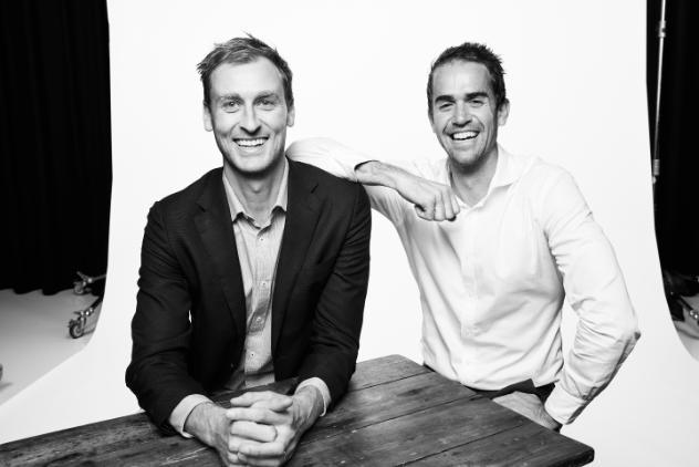 Two young men in corporate attire sitting. The man on the left has his elbow on the other mans should, both men are smiling. The image is in black and white.