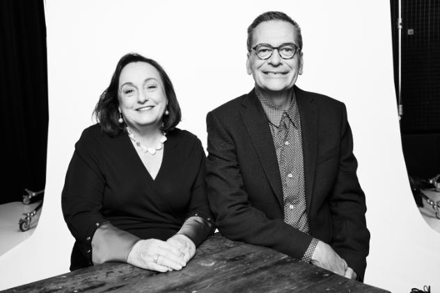 Black and white photo of a couple sitting together at a table. The woman has short hair and has a hands together on the table. The man is sitting beside her with his right elbow resting on the table. He has short dark hair and glasses.