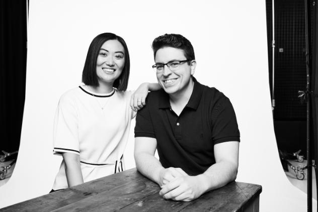 A young female and male are sitting at a table looking at the camera smiling. The female has her elbow on the males shoulder. He is leaning with both elbows on the table. This image is in black and white.