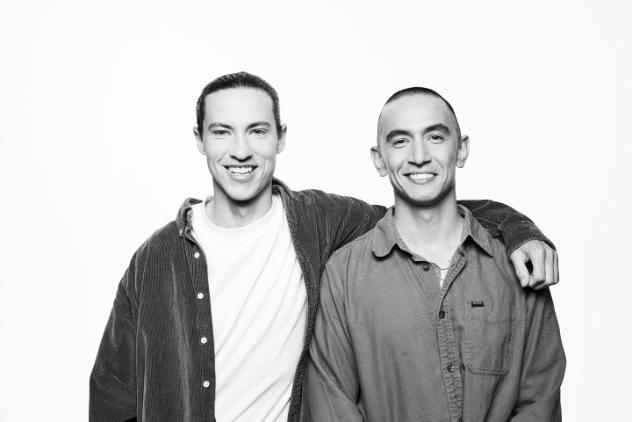 Two young men standing next to each other, smiling, looking directly at the camera. The man on the left has his arm around the other mans shoulders. This image is in black and white.