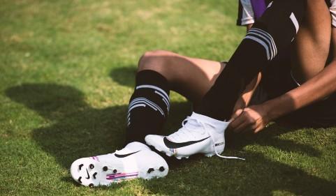 View of male sitting on grass, putting on white football boots, and wearing black socks. Image is of legs and boots only.