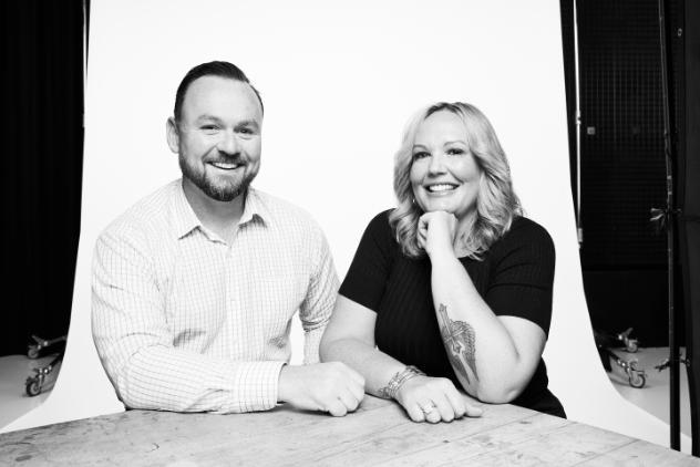 Two friends sitting together laughing. Male friend with facial hair is on the left in a square checkered top the female on the right has her elbow on his shoulder and is laughing and wearing all black. The image is in black and white.