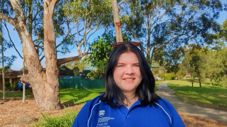 female student in blue t-shirt smiling at camera with trees and greenery in the background.