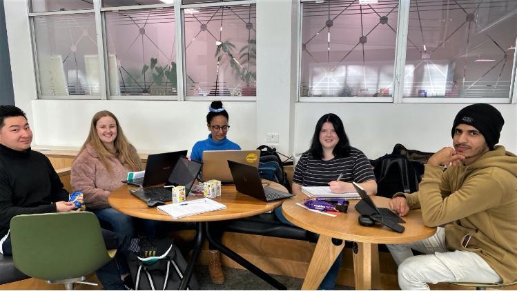 group of 5 students sitting at tables with laptops smiling at camera