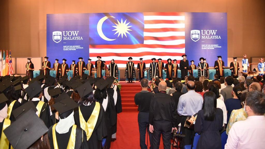 Back of audience heads looking at 2022 UOW Graduation ceremony with Malaysian flag in background