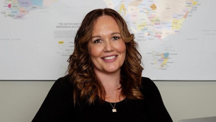 A woman smiling at the camera. Behind the womans head there is a map on the wall of the different Indigenous Nations of Australia.