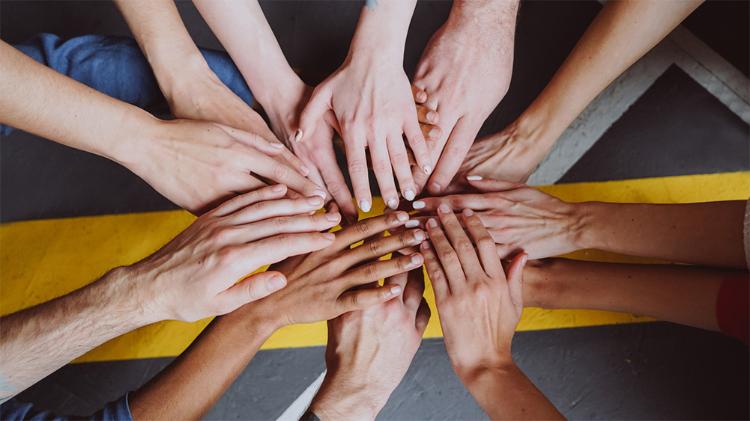 A diverse group of hands stacked together in a circle on a gray floor, symbolizing unity and collaboration.