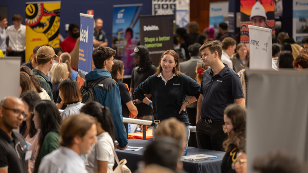A bustling career fair with people interacting at various booths. Two representatives in navy shirts engage with attendees, creating a lively, professional atmosphere.