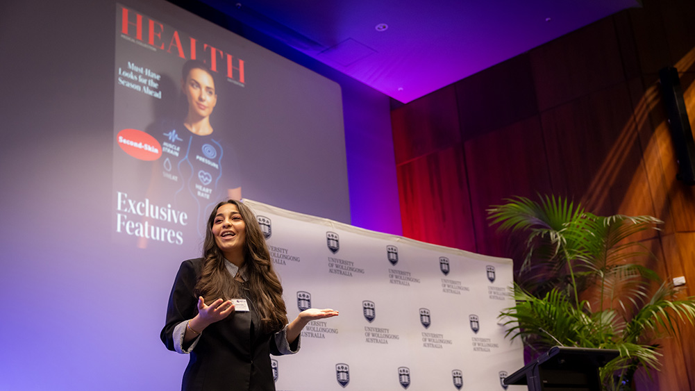 A university student presents on stage at the UOW Three Minute Thesis Final, speaking in front of a projected slide and a University of Wollongong backdrop in an auditorium with purple lighting and indoor plants.