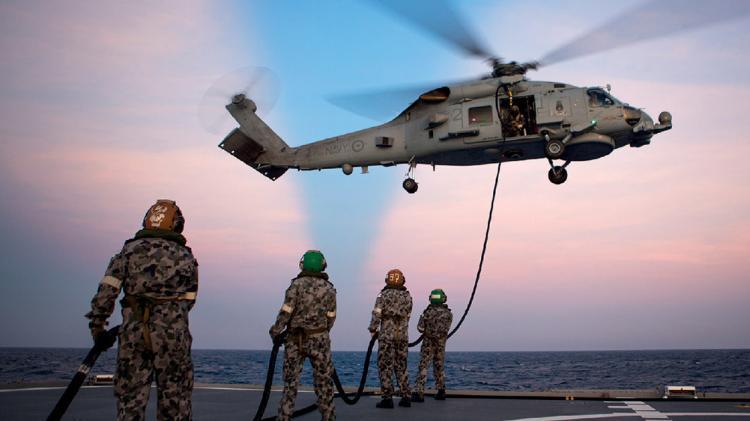 Military helicopter landing on the ship at dusk. Defence personnel stand below with ropes.