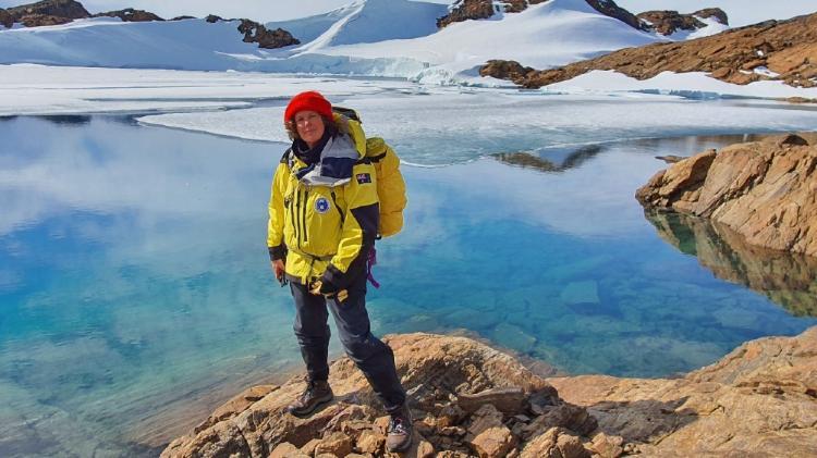 Person wearing a yellow weather coat and red beanie standing on brown rocks on a water edge with an Antarctic scenery
