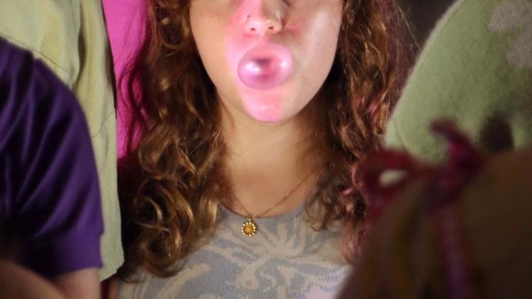 Person with curly brown hair wearing a gold chain and pendant is blowing a bubble with pink gum.