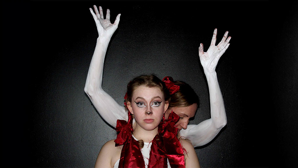 A woman stands infront of someone who has their hands in the air, the arms and hands are painted white. The woman at the front is in plaited pig tails with red satin bows. she has face paint on her nose and under her eyes to make her look like a dear.