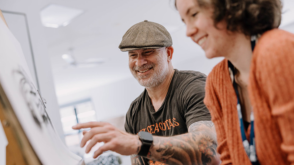 Two people participating in an art activity in a classroom or studio setting. One person wearing a flat cap and a dark graphic T-shirt gestures toward a drawing or artwork displayed on an easel, while another person sits nearby. The scene includes art materials, bright natural light and a creative learning environment focused on visual arts and collaboration.