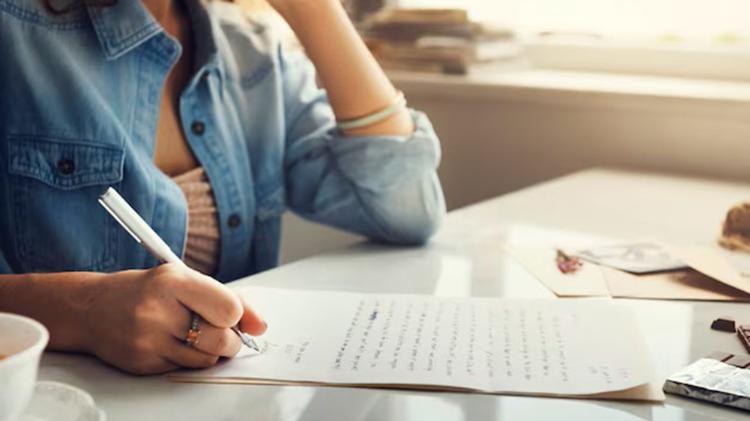 A woman in a denim shirt writes a letter on a table, with a cup and scattered photographs visible in soft, natural light.