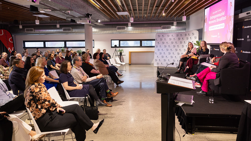 Audience seated in a modern conference room listens attentively to a panel of four speakers on stage during an 