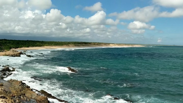 A panoramic view of a tranquil beach with turquoise waves lapping against rocky shorelines, under a partly cloudy blue sky.