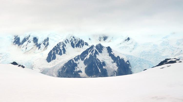 Snow-covered mountains with jagged peaks rise in the background under a cloudy sky, partially obscured by mist. The foreground shows a flat, snowy expanse.