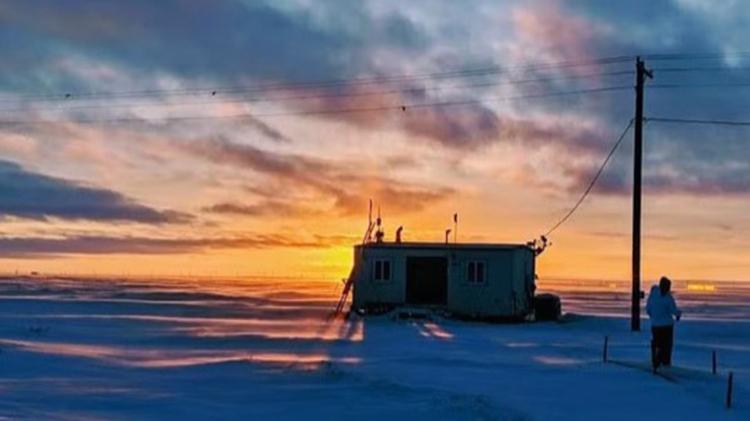 A person stands near a small, isolated building in a snowy landscape at sunset, with power lines above and a colorful, cloud-filled sky in the background.
