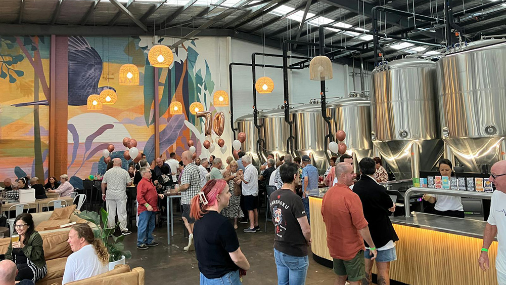 A lively gathering inside a brewery with guests mingling around shiny silver brewing tanks and colorful wall art, illuminated by pendant lights.