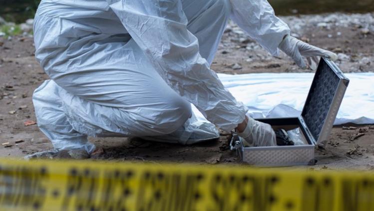 People wearing a white forensic suit is crouched down putting something in a small steel box. Yellow tape with the words 