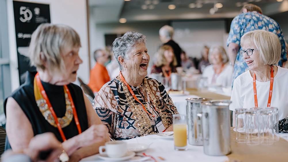 A group of elderly women with lanyards enjoy a lively conversation at a gathering. They are seated at a table with cups, a juice glass, and pitchers.