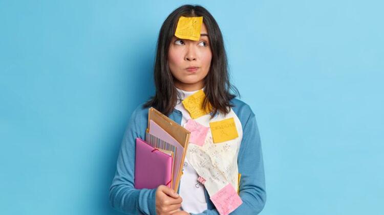 A woman holding several books in front of a sky blue coloured wall. Coloured postnotes are on her including her forehead. She is wearing a blue jumper and has a questioning expression on her face.