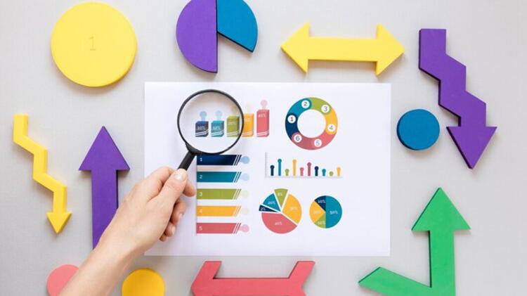 A hand holds a magnifying glass over colorful graphs and charts surrounded by various geometric shapes on a grey background.