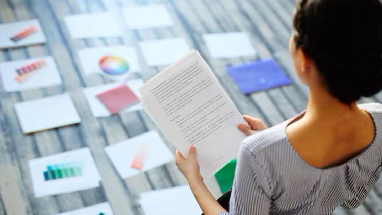 A woman reads a document while seated among colorful design printouts on a wooden floor, deep in thought.