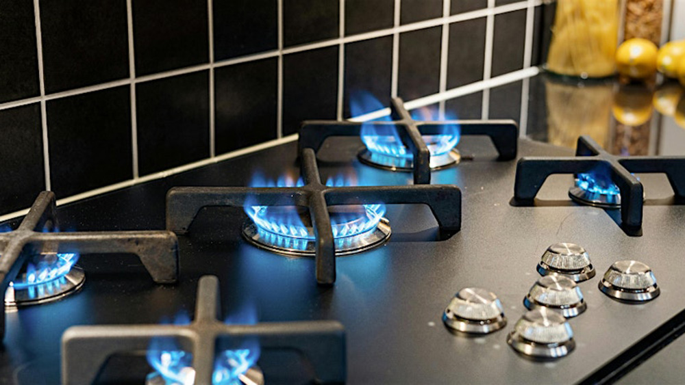 A gas stove with four burners lit with blue flames, black grates, and control knobs on a black countertop, set against a tiled backsplash.