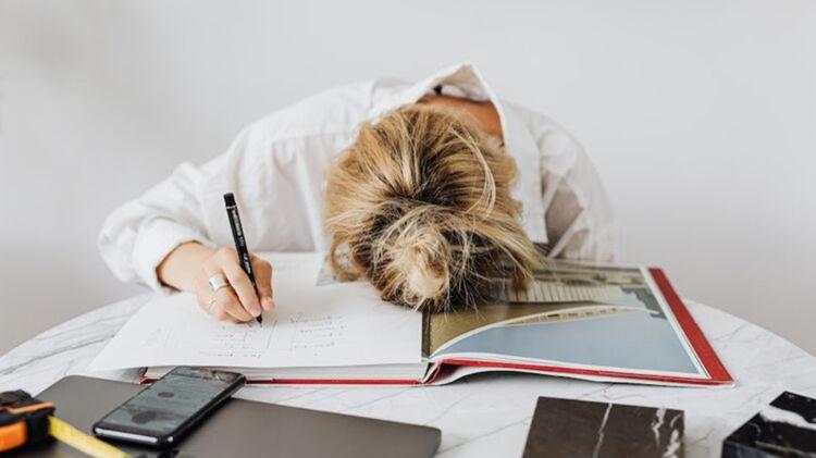 A woman rests her head on an open design book, overwhelmed, with a pen in hand and a smartphone nearby on a table.