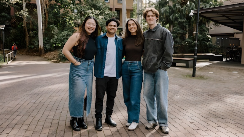 Four students stand facing camera and smiling. They are on a paved area with trees in the background