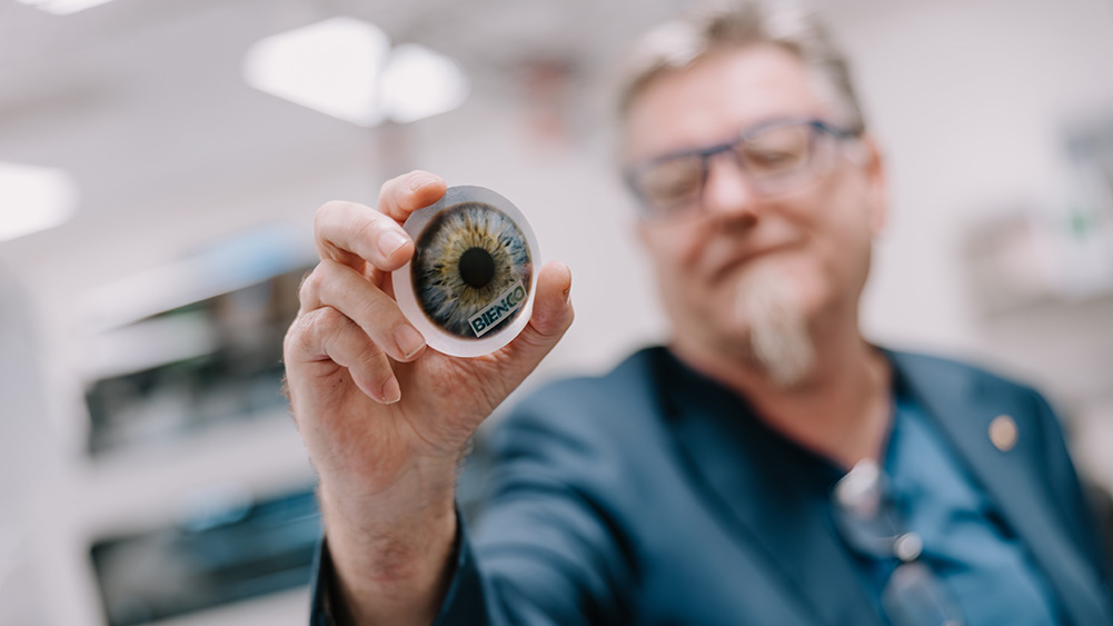 Close-up photograph of a person in a laboratory or research setting holding a circular object toward the camera. The object features a detailed image of a human iris and pupil, with the word “BIENCO” visible near the bottom edge. The camera focuses sharply on the object and the person's hand, while the person and the background are softly blurred. The background contains bright indoor lighting